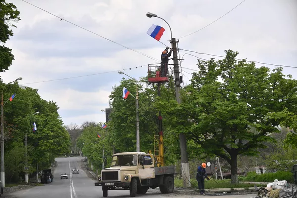 Кормандони ширкати шабакаи рӯшноии берунии "Горсвет" парчами Русияро дар симчӯби чароғдоре дар Мариупол меовезанд. Кормандони ширкати шабакаи рӯшноии берунии "Горсвет" парчами Русияро дар симчӯби чароғдоре дар Мариупол меовезанд. - Sputnik Тоҷикистон