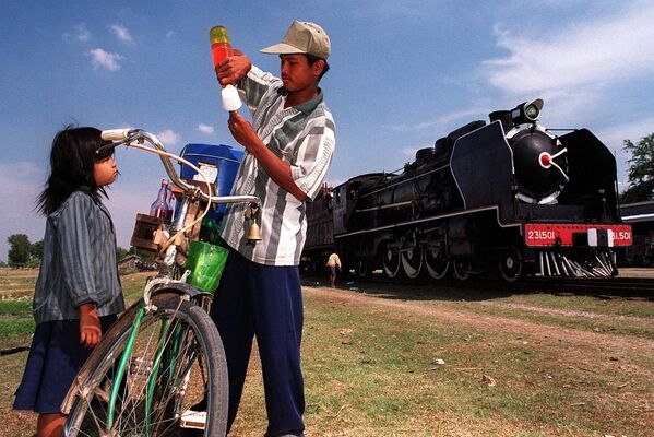 Камбоджиец готовит мороженое для маленькой девочки. За ними - старый паровоз &quot;Пасифик 231&quot; останавливается на небольшой станции в 50 км к югу от Пномпеня, 2000 год. - Sputnik Таджикистан