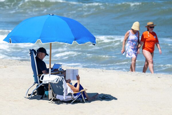 Посетители пляжа идут вдоль воды, пока президент США Джо Байден (слева) и первая леди США Джилл Байден сидят под зонтиком в Рехобот-Бич, Делавэр. - Sputnik Таджикистан