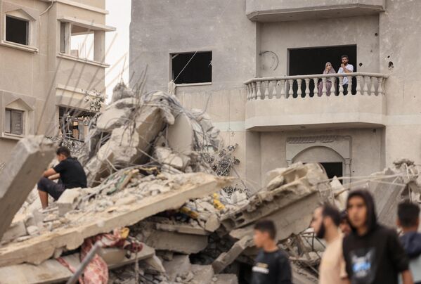 Пара наблюдает с балкона поврежденного дома, как люди осматривают ущерб, нанесенный израильской бомбардировкой в Хан-Юнисе на юге сектора Газа 12 ноября 2023 года. - Sputnik Таджикистан