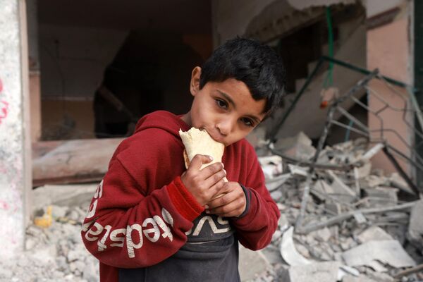 Ребенок ест сэндвич, стоя перед зданием, поврежденным в результате израильской бомбардировки на юге сектора Газа. - Sputnik Таджикистан