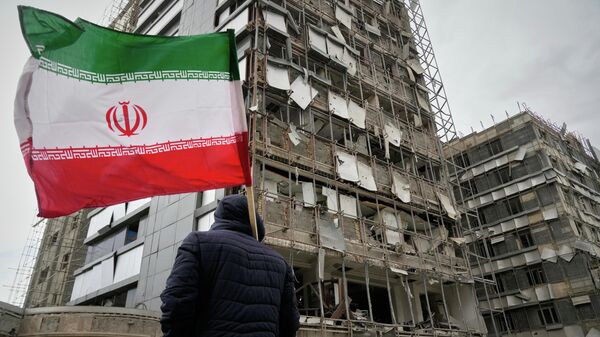 Мужчина держит иранский флаг, осматривая поврежденный фасад больницы имени Ганди в Тегеране, Иран. - Sputnik Тоҷикистон