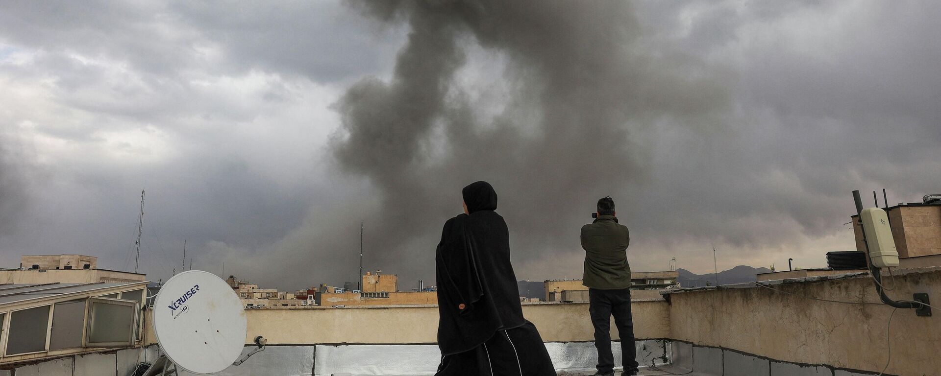Люди наблюдают за поднимающимся дымом после взрыва, произошедшего после ударов Израиля и США по Ирану  2 марта 2026 года.  - Sputnik Тоҷикистон, 1920, 02.03.2026