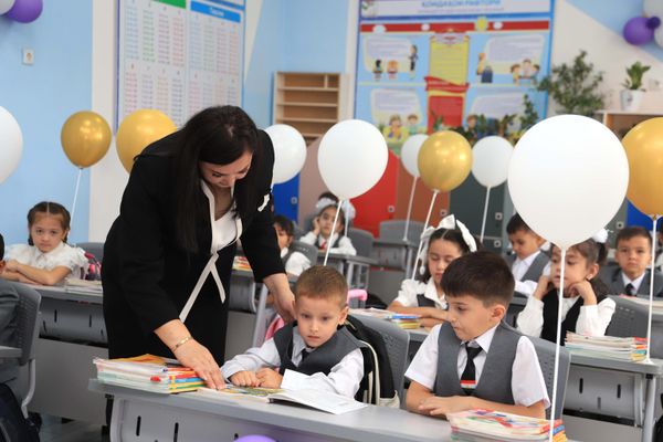 Учительница наклонилась к ученикам за партой и показывает что-то в учебнике. - Sputnik Таджикистан
