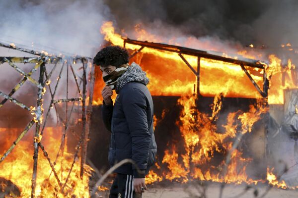 Мигрант идет мимо горящей конструкции во время демонтажа лагеря мигрантов в Кале (Франция) - Sputnik Таджикистан